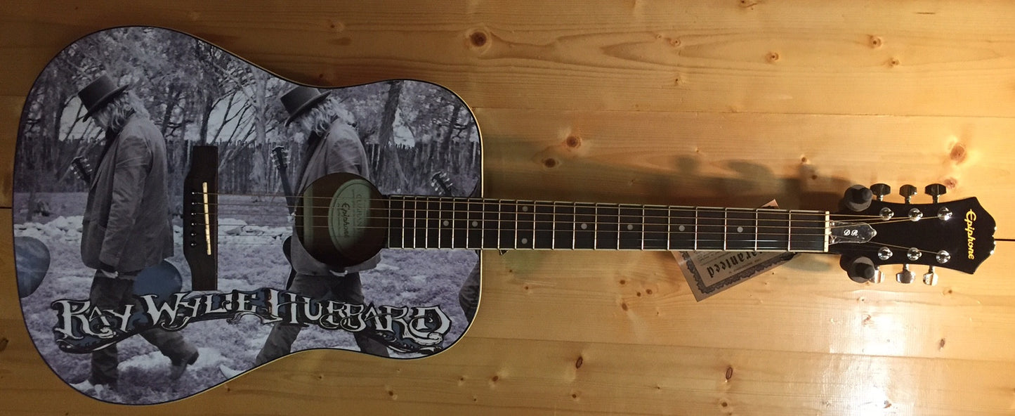 Acoustic guitar with a custom black-and-white graphic on the body featuring two figures walking outdoors and the text ‘Ray Wylie Hubbard.’ The guitar has a natural wood neck and headstock with Epiphone branding.