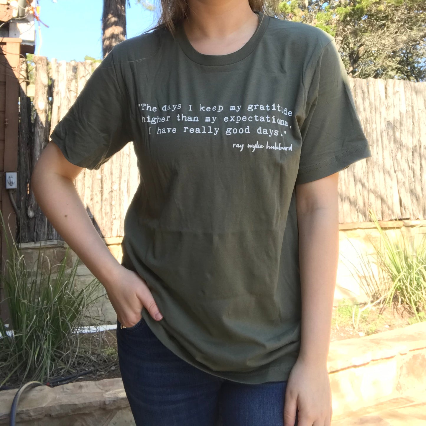 A person wearing a green unisex t-shirt with text on it, standing outdoors.