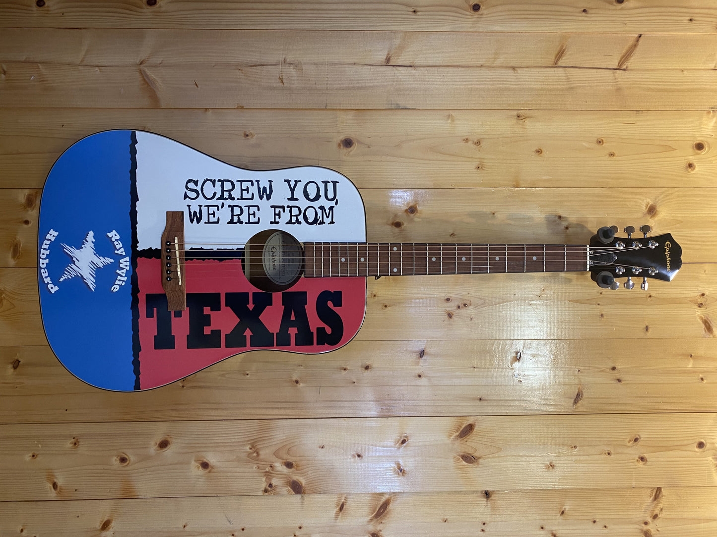 An acoustic Epiphone guitar with a Texas flag-themed design, featuring text 'Screw You We're From Texas' on the body, laid on a wooden surface.