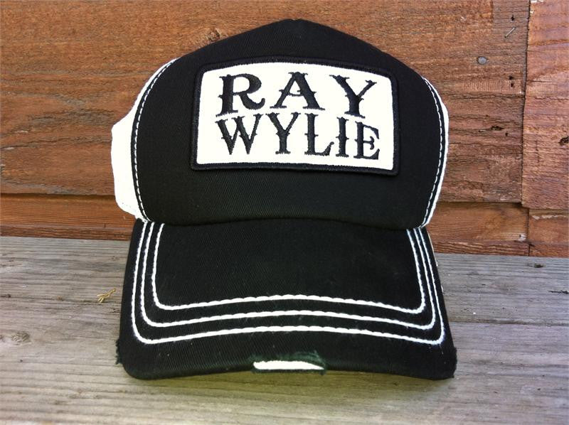 A black and white trucker hat with 'RAY WYLIE' text on the front panel, placed on a wooden surface.